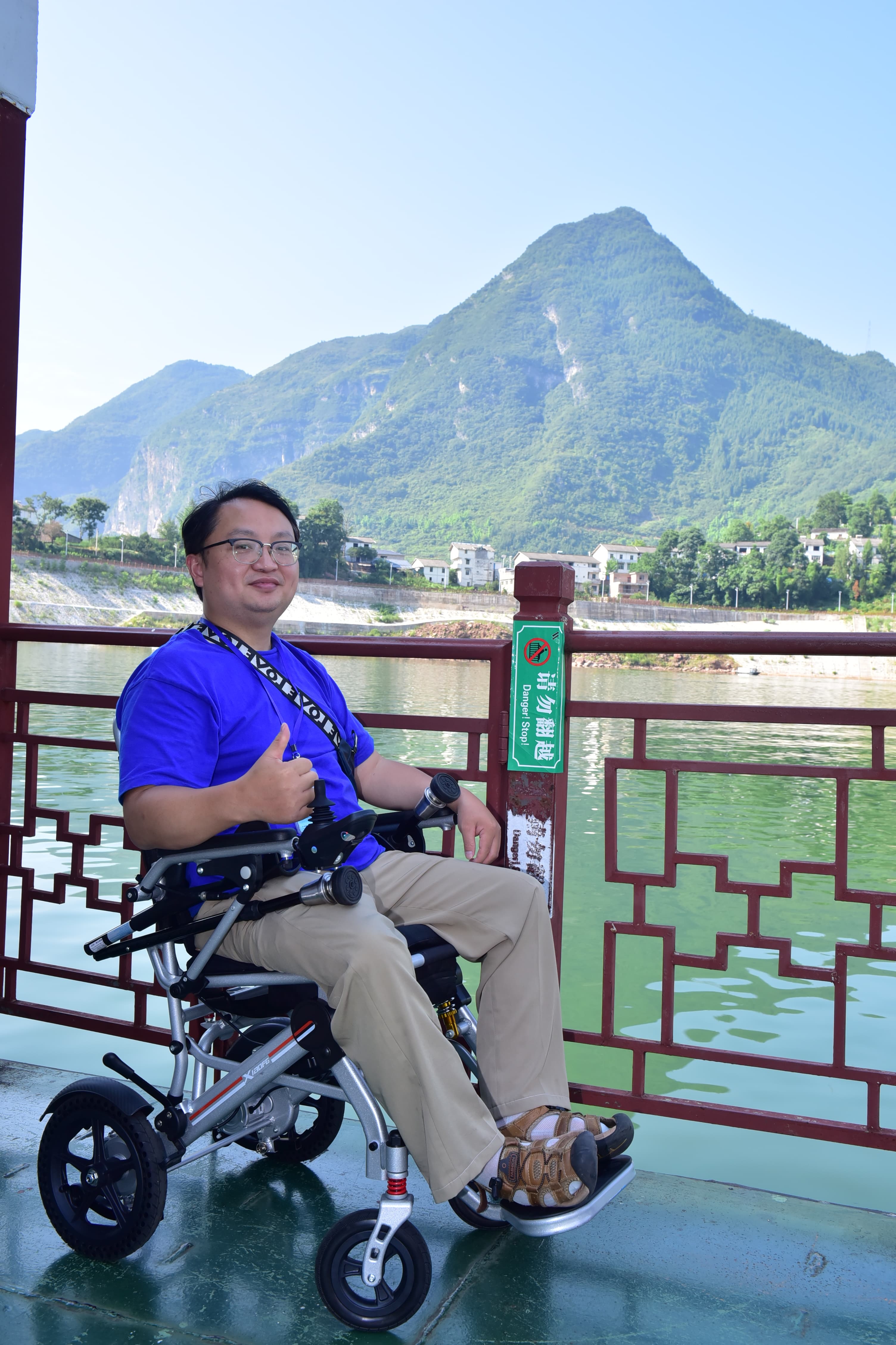 Xiong Ge, founder of AITrans.Video, seated in his power wheelchair and giving a thumbs-up to the camera, with a river and green mountains behind him